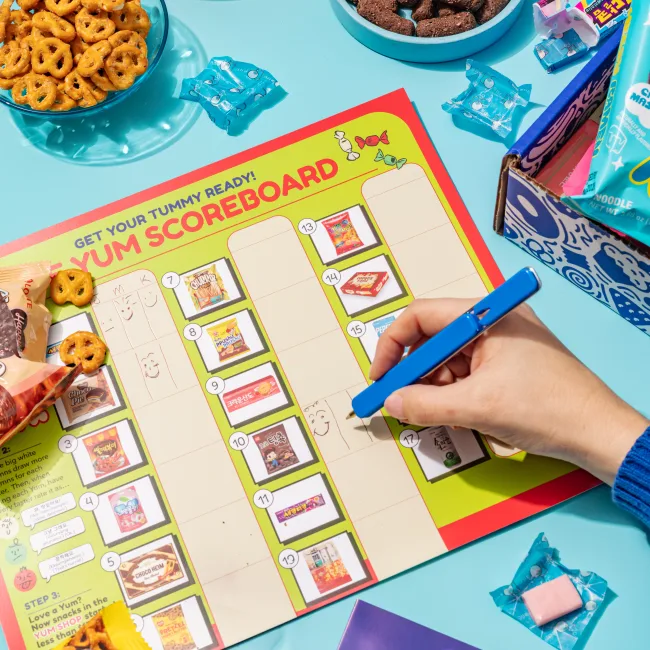 A hand filling in the Yum Scoreboard next to a box of snacks.